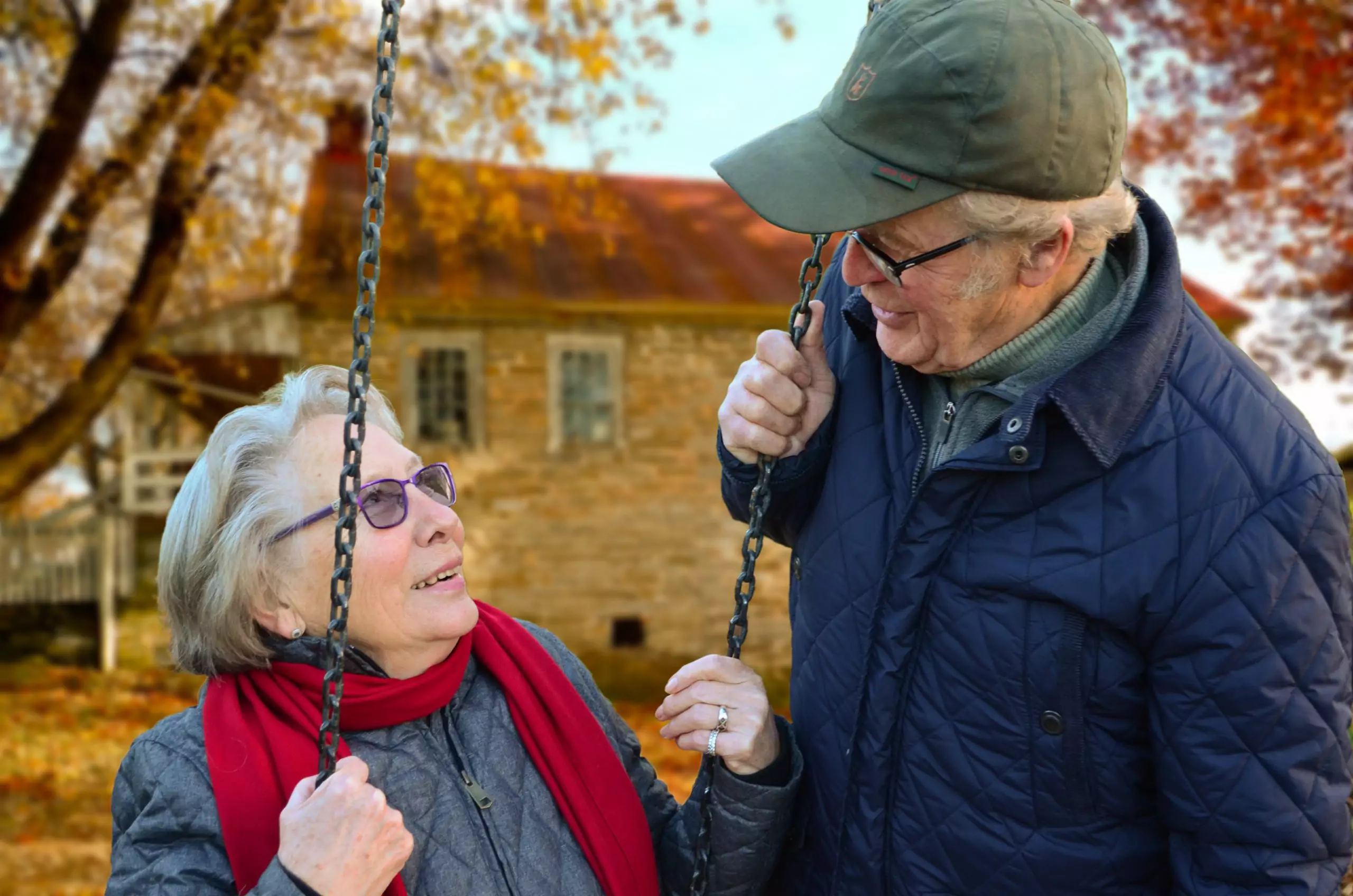 An elderly couple smile at one another outside while the woman sits on a swing
